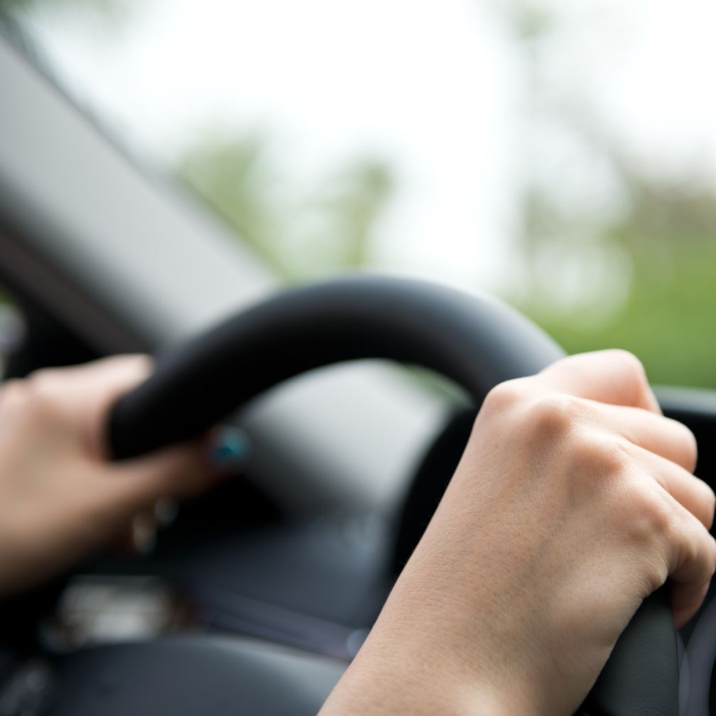 Close-up of two hands gripping a steering wheel inside a car, with a blurred view of greenery and the dashboard visible in the background.