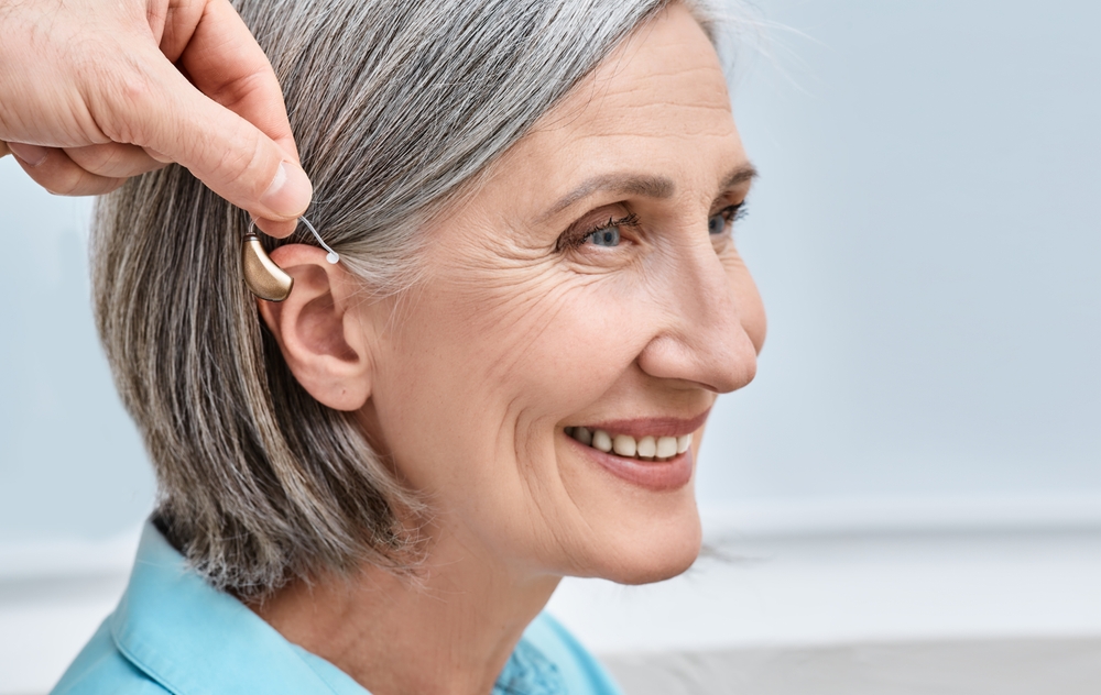 A smiling older woman with gray hair has a hearing aid placed behind her ear by another persons hand.