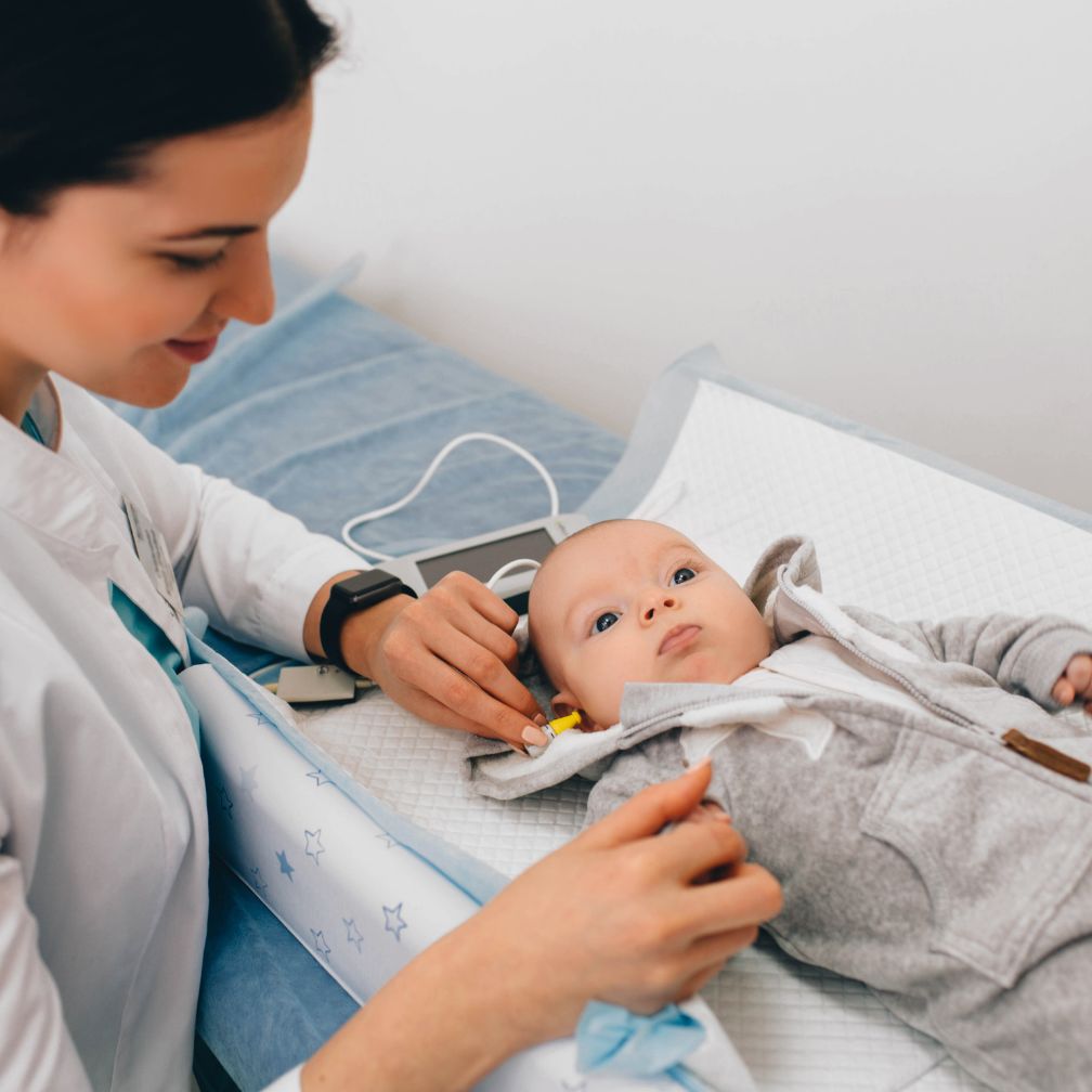 A healthcare professional performs a hearing test on a baby lying on an exam table, using small devices placed in the infants ears. The baby looks up while the professional gently holds the babys hand.