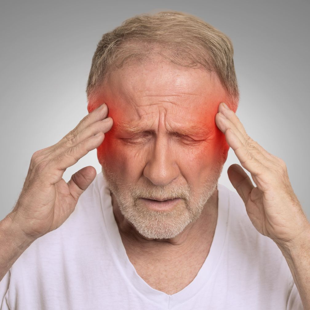 An older man holds his head with both hands, eyes closed in pain. Red shading highlights his temples, indicating a headache or migraine.