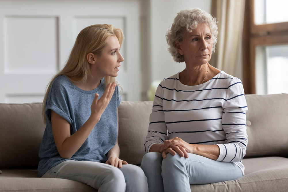 A young woman sits on a couch, gesturing and talking to an older woman with gray hair who looks away, appearing upset or thoughtful. Both women have serious expressions in a home setting.