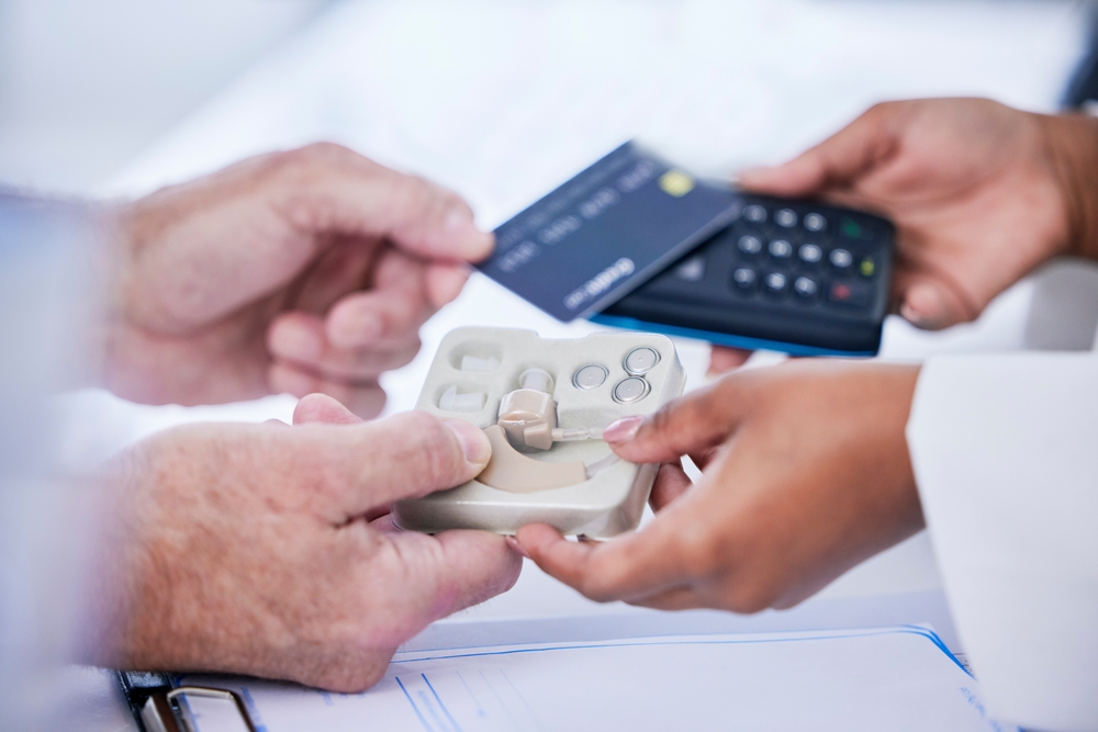A person makes a payment with a credit card and card reader while receiving a hearing aid from another person, with paperwork visible on the table.