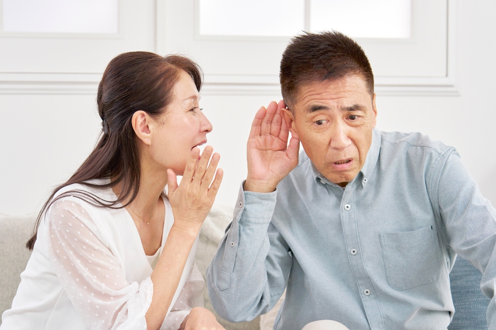 A woman leans in to whisper to a man who cups his ear and looks confused or surprised, both sitting on a couch in a bright room.