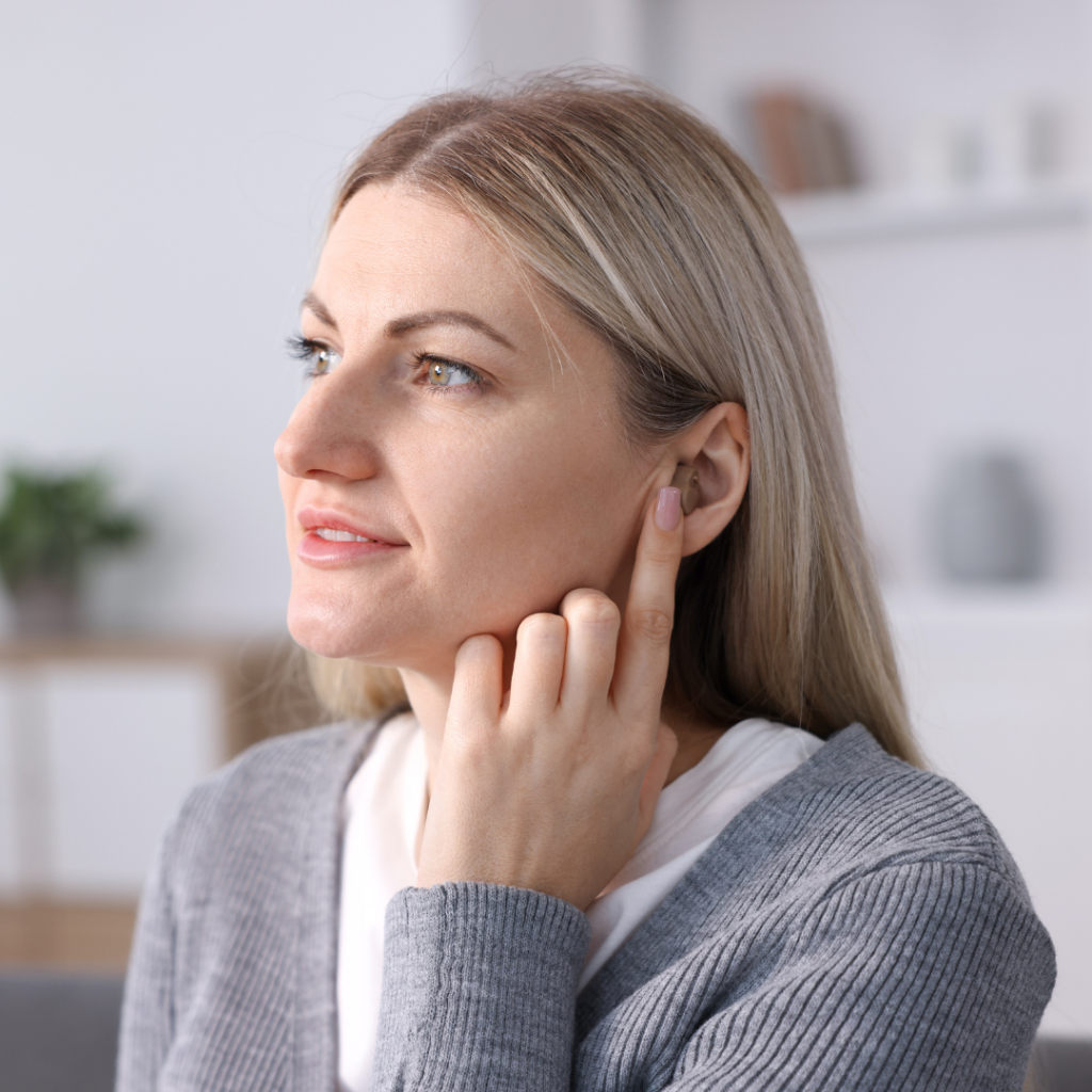 A woman with blonde hair touches her ear, appearing to listen closely. She is indoors, wearing a gray sweater, and looking slightly to the side with a thoughtful expression. The background is softly blurred.