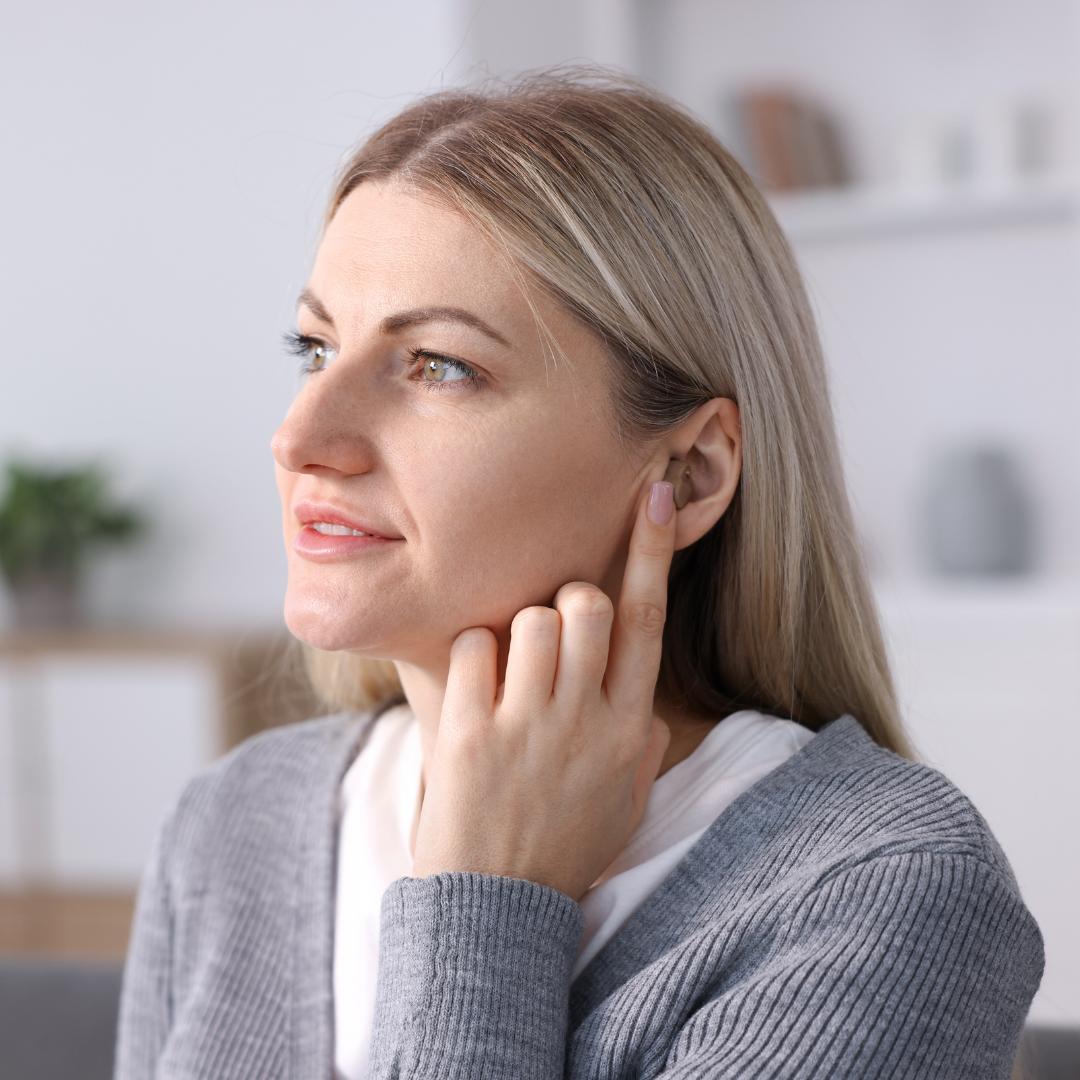 A woman with blonde hair touches her ear, appearing to listen closely. She is indoors, wearing a gray sweater, and looking slightly to the side with a thoughtful expression. The background is softly blurred.